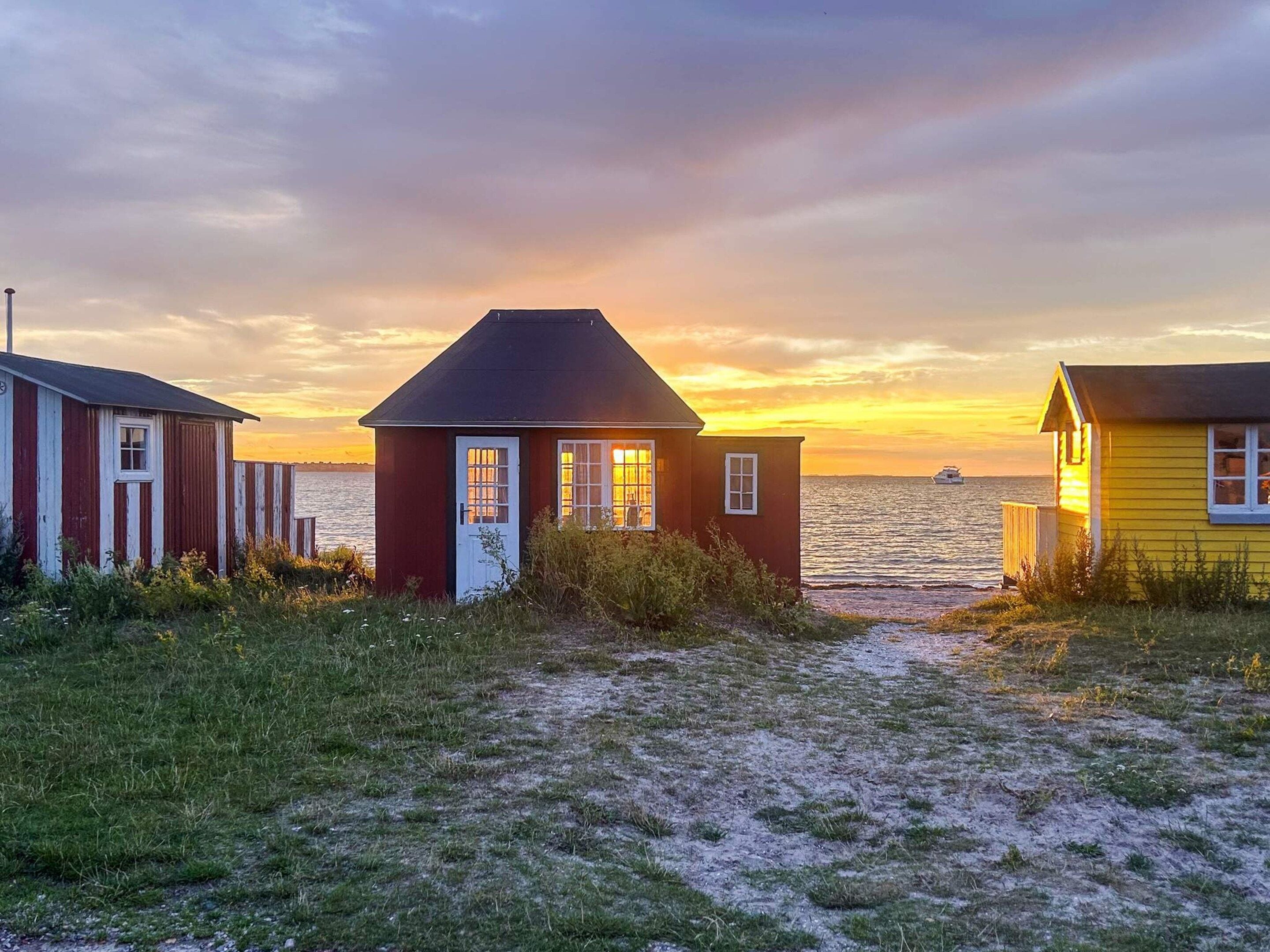 Une scène côtière avec trois petites maisons en bois, dont une rouge et deux jaunes, offrant une vue sur la mer et un coucher de soleil en arrière-plan.