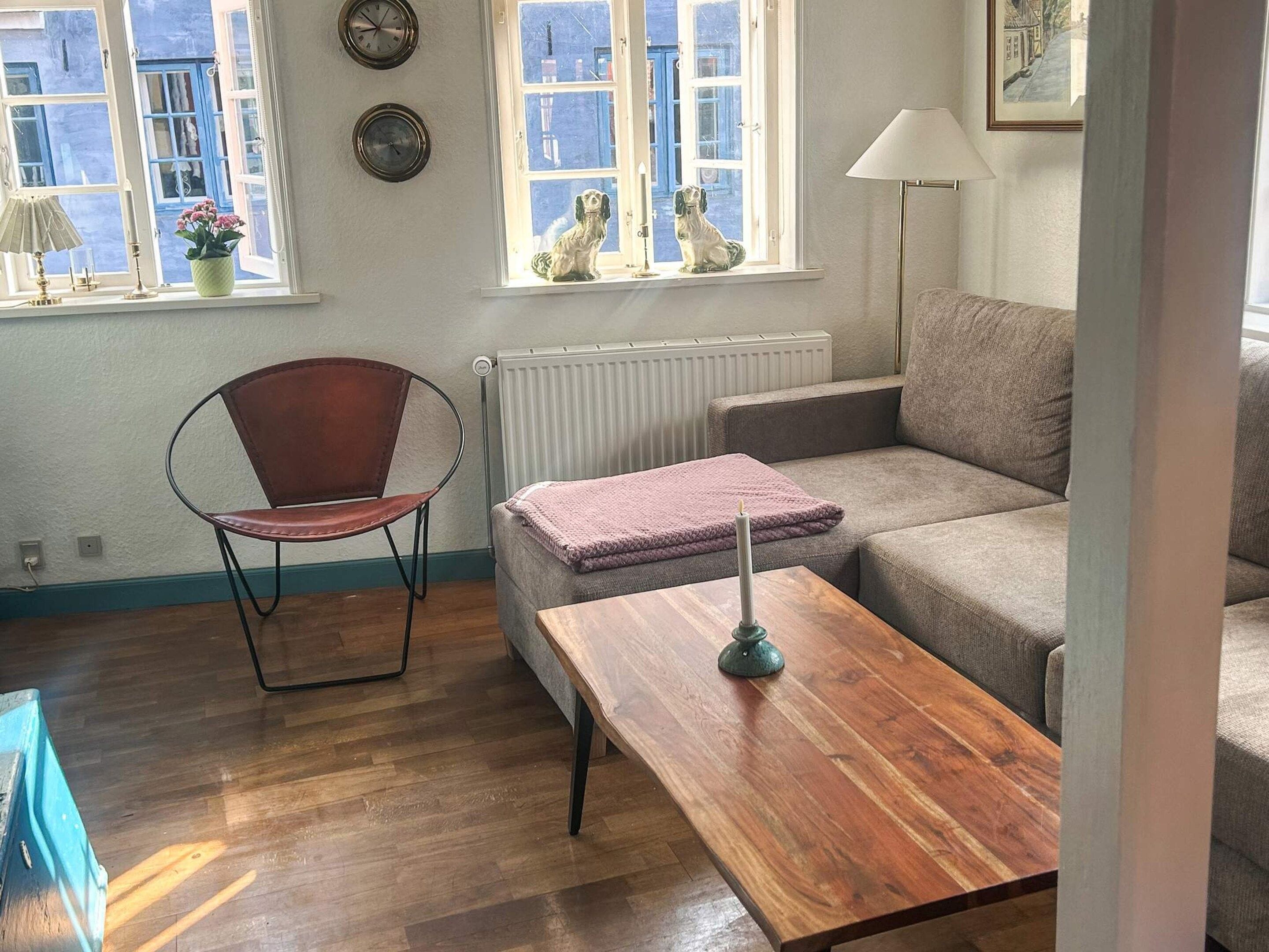 Un salón con suelo de madera, un sillón de cuero marrón, un sofá gris, una mesa de centro de madera, un radiador y una ventana con vistas a edificios.