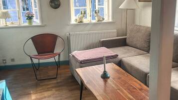 Un salón con suelo de madera, un sillón de cuero marrón, un sofá gris, una mesa de centro de madera, un radiador y una ventana con vistas a edificios.