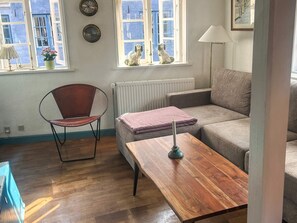 En stue med trægulv, en læderlænestol i brunt, en grå sofa, et træbord og en radiator. Et vindue viser udsigt til bygninger.