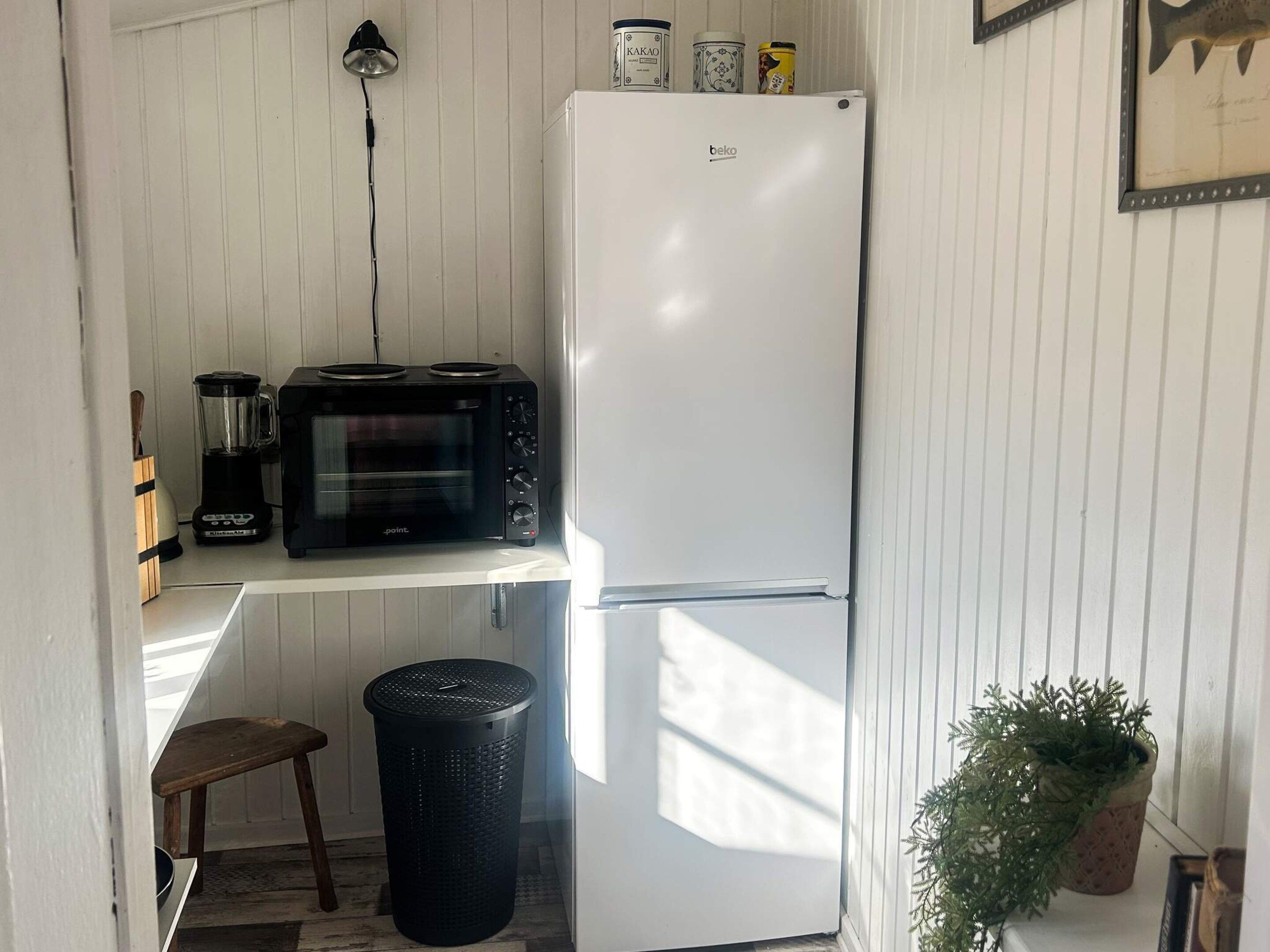 A compact kitchen with a white refrigerator, a black microwave, and a blender on a white countertop. There is a trash bin and a potted plant in the corner.
