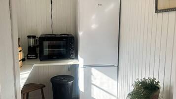 A compact kitchen with a white refrigerator, a black microwave, and a blender on a white countertop. There is a trash bin and a potted plant in the corner.