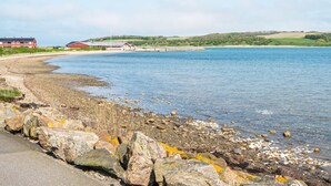 Paesaggio costiero con spiaggia di ciottoli, cielo sereno e edifici in lontananza.