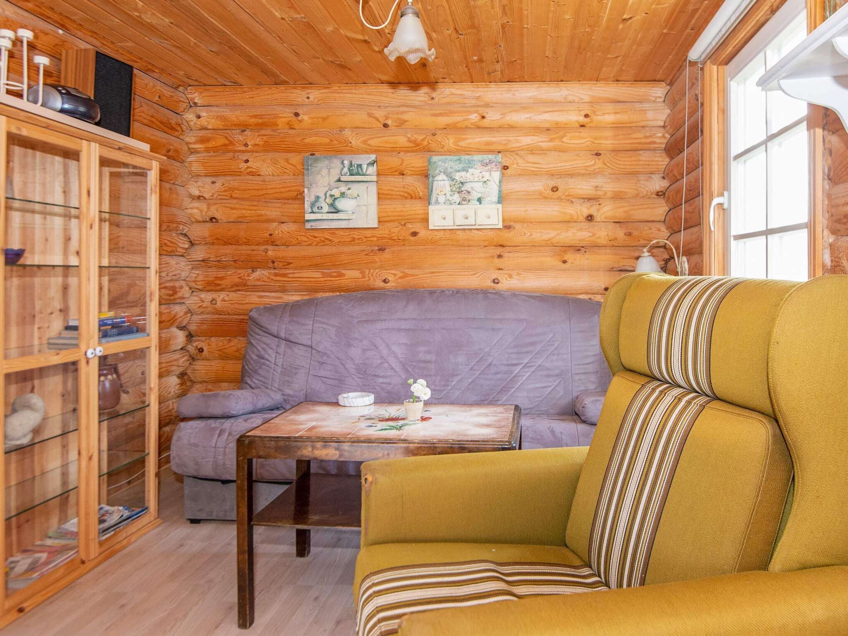 A cozy cabin interior with a wooden log wall, a purple sofa, a yellow armchair, and a small table with a teapot and cup.