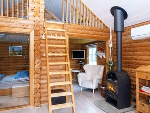 A log cabin interior with a wooden ladder, a white armchair, a fireplace, and a television.