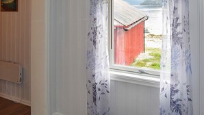 A room with a wooden floor, a window with a view of a red building and a body of water, and a table with a book. - Holiday Home in MÃ¥ndalen (Rauma)