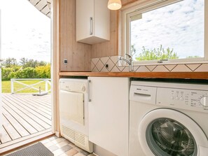 A laundry room with a washing machine and dryer, wooden cabinets, and a window overlooking a garden.