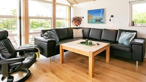 A modern living room with a large black sectional sofa, a wooden coffee table, and a view of greenery through large windows. - 5 Star Holiday Home in Lemvig (Lemvig)