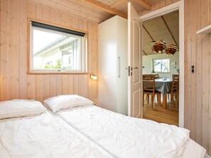 A room with a bed, a window, a door leading to a dining area, and a ceiling with wooden beams.