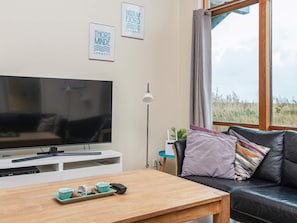 A modern living room with a flat-screen TV, a wooden coffee table with a remote and a small dish, a black leather sofa with colorful cushions, and framed artwork on the wall.