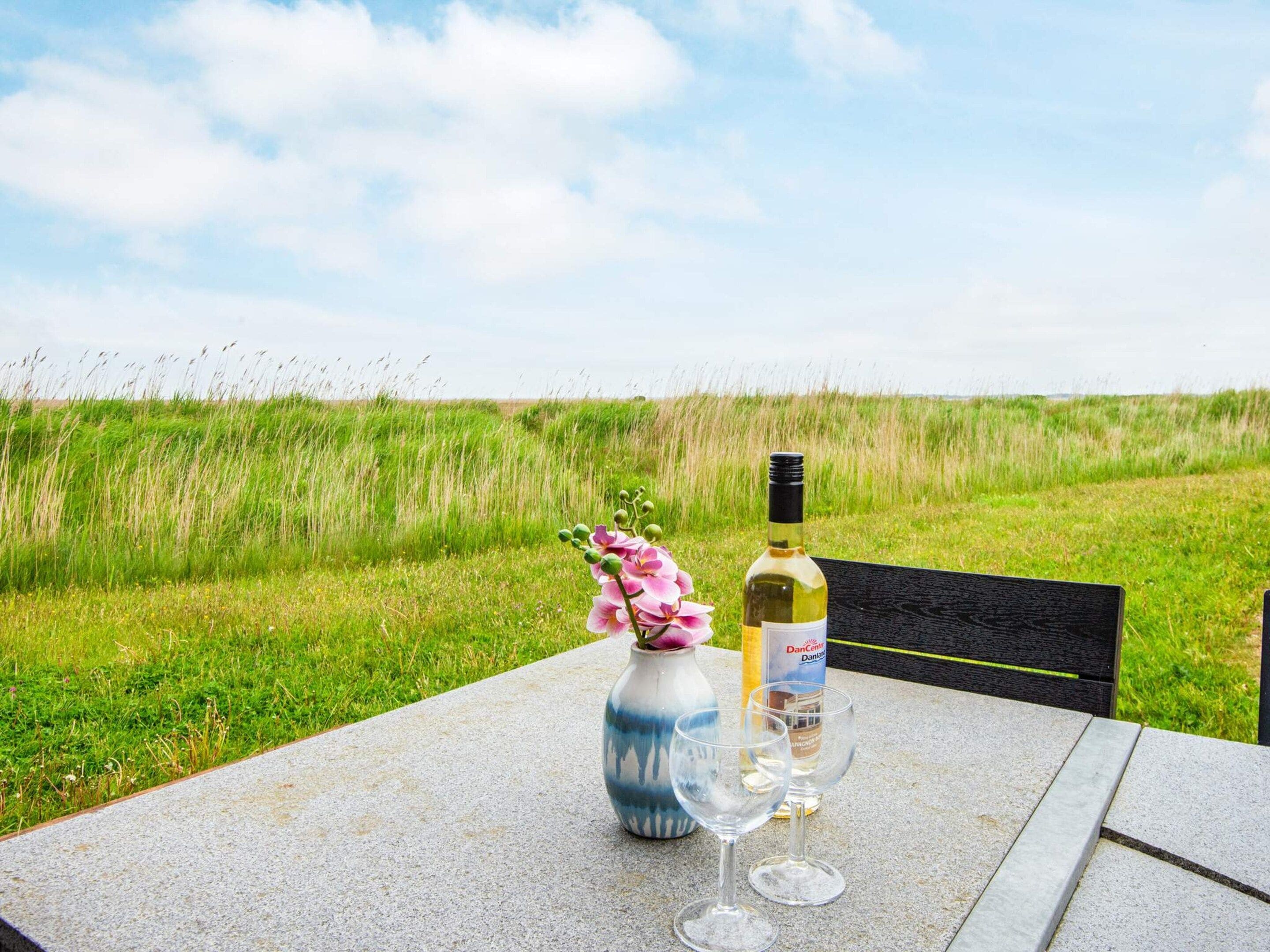 A table with a bottle of wine, two glasses, and a vase with flowers, set against a grassy field.