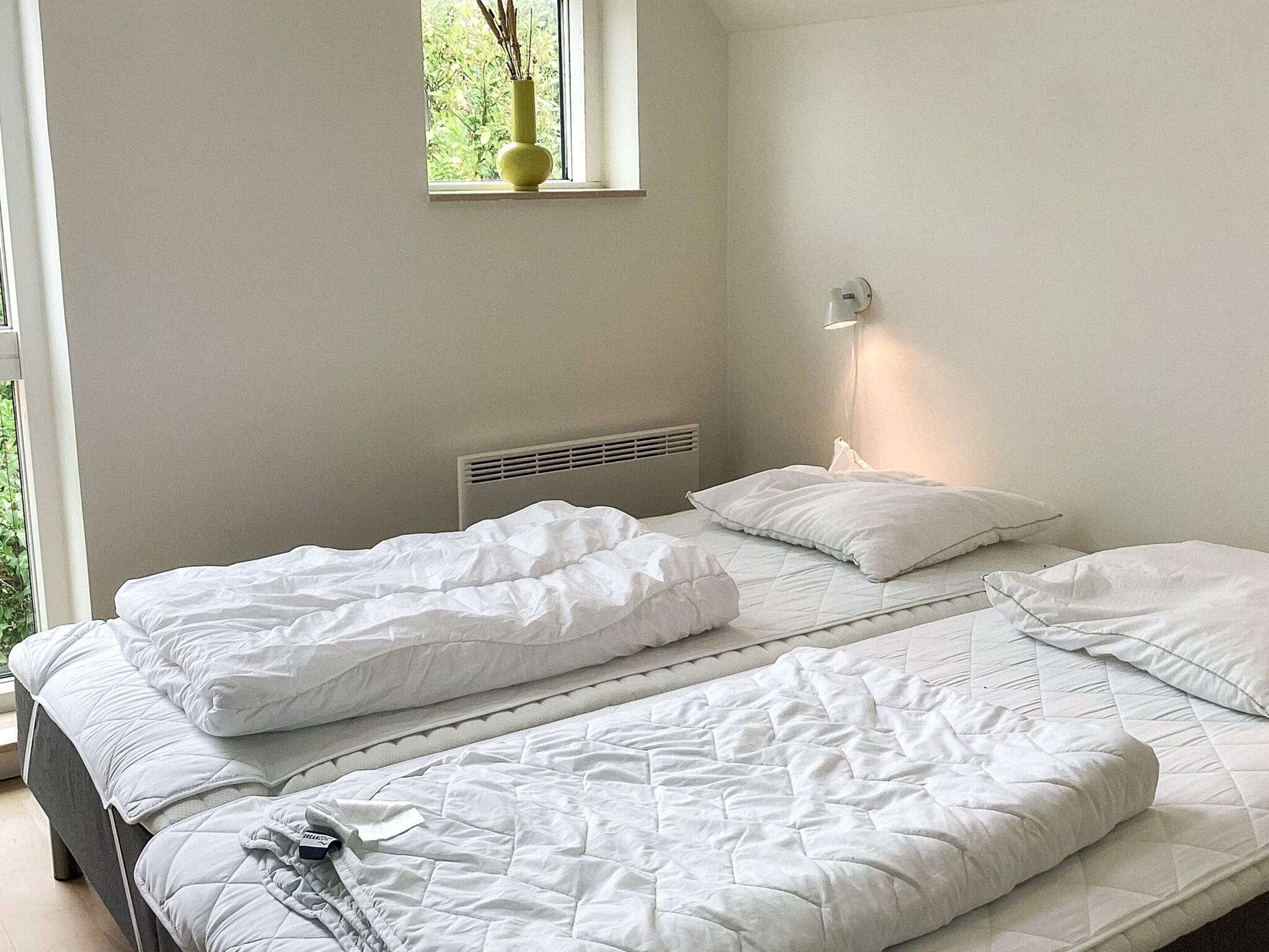 A bedroom with two beds, a window with a plant, a wall-mounted light, and a radiator.