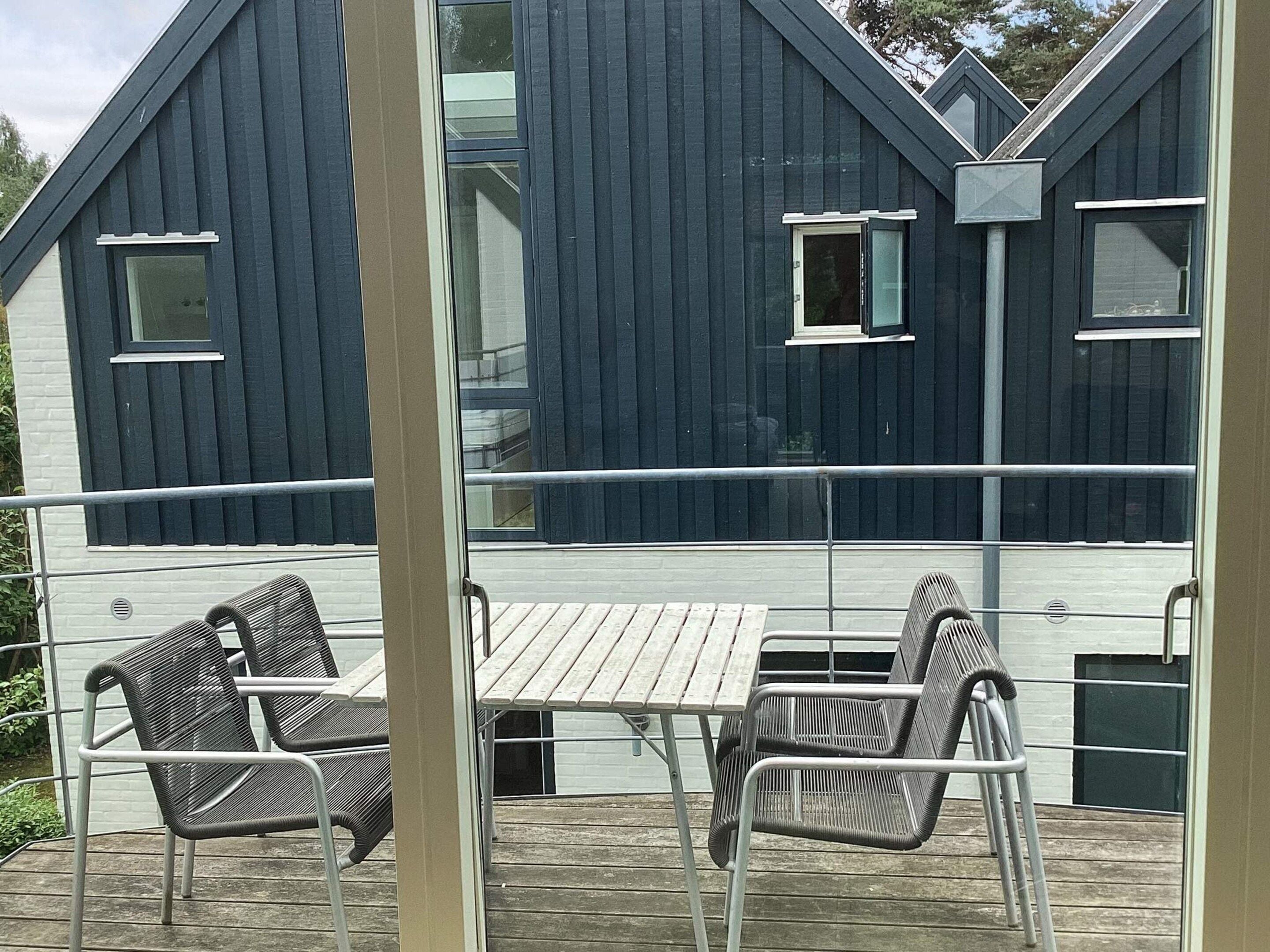 A wooden deck with metal chairs and a table, overlooking a dark-colored building with windows and a balcony railing.