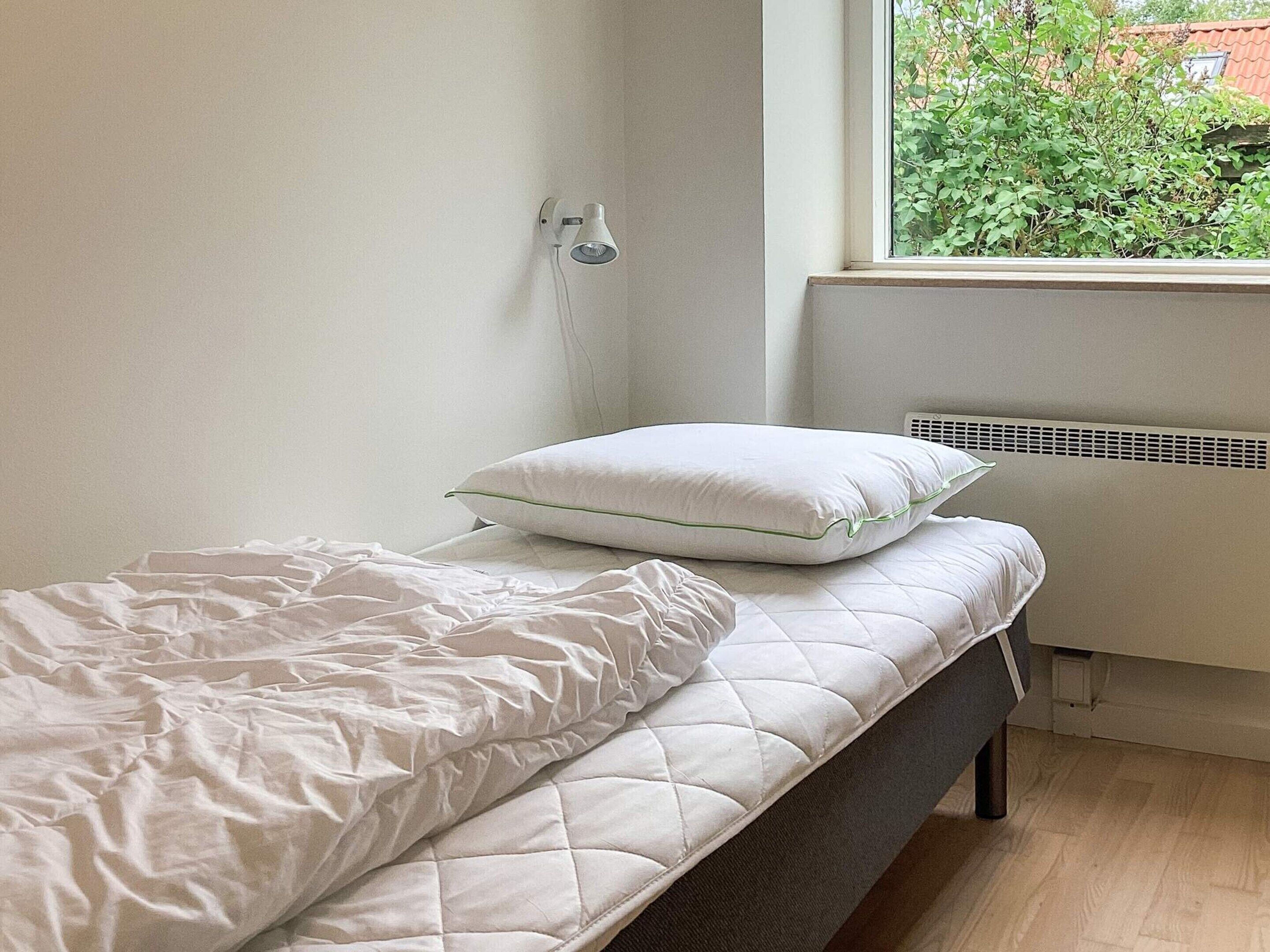 A small, neatly made bed with a white quilted mattress and a pillow, a white wall, and a window with a view of greenery outside.