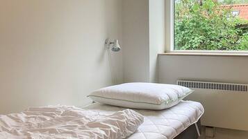 A small, neatly made bed with a white quilted mattress and a pillow, a white wall, and a window with a view of greenery outside.