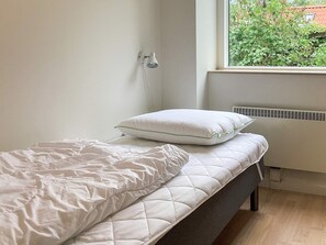 Un petit lit bien fait, avec un matelas matelassé blanc et un oreiller, un mur blanc et une fenêtre donnant sur un paysage verdoyant.
