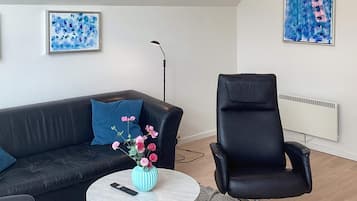 A modern living room with a black leather sofa, a round coffee table with a vase of flowers, a black office chair, and two abstract paintings on the walls.