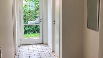 A narrow hallway with a white door leading to a garden. The floor is tiled, and there is a wall-mounted electrical box on the right.
