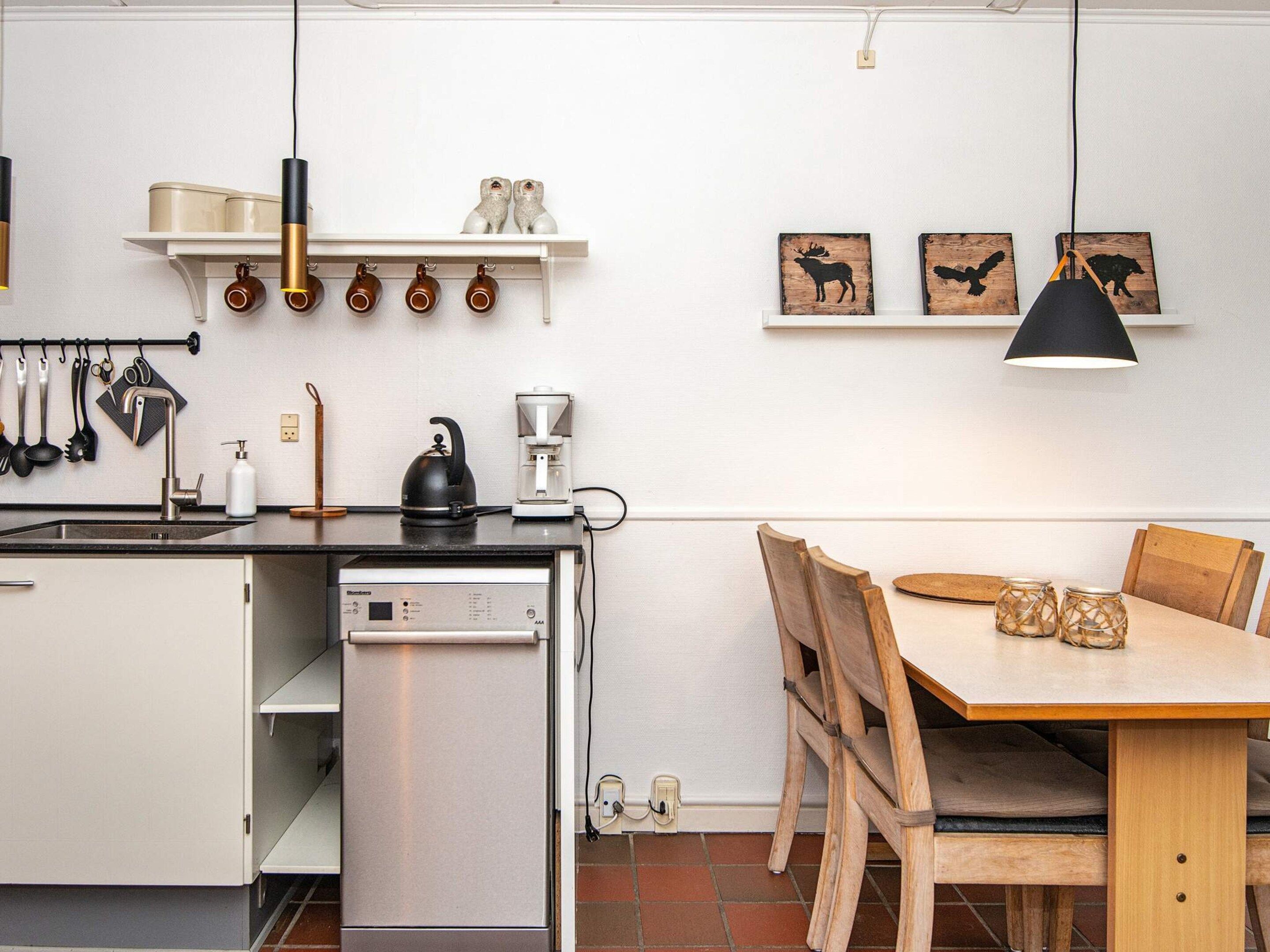Cocina compacta con lavavajillas, hervidor, cafetera y utensilios colgantes. Zona de comedor con mesa y sillas de madera.