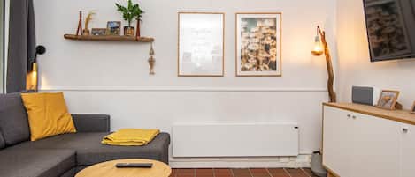 A modern living room with a grey sofa, a small round table, a radiator, a TV mounted on the wall, and a shelf with decorative items.
