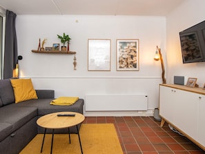 A modern living room with a grey sofa, a small round table, a radiator, a TV mounted on the wall, and a shelf with decorative items.