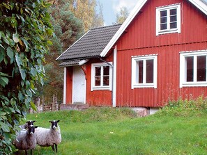 Une maison rouge aux contours blancs, dotée de nombreuses fenêtres, entourée d’une pelouse verdoyante et d’arbres.