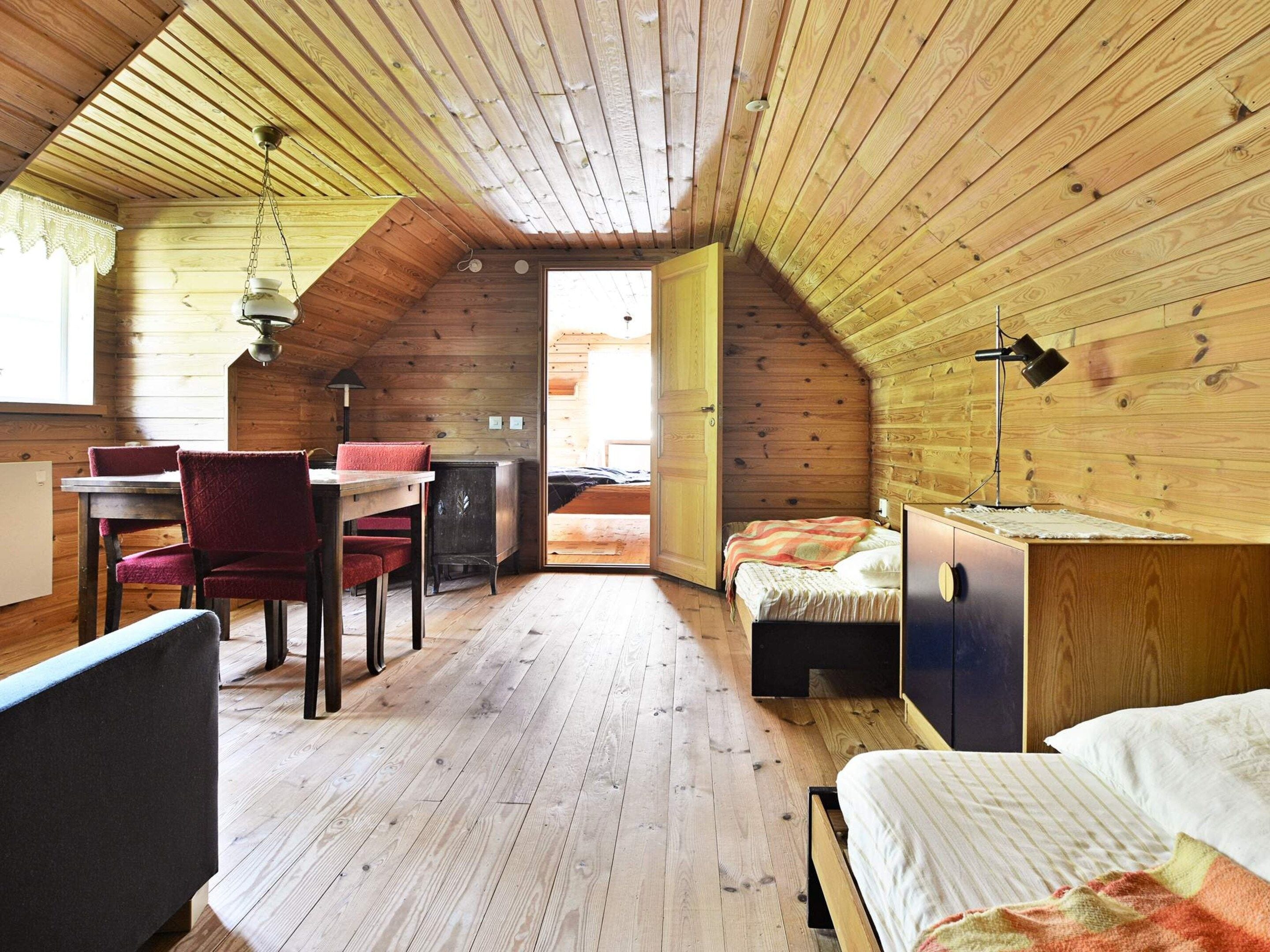 A cozy attic room with a wooden ceiling, a dining table with chairs, a bed, and a desk with a lamp.