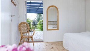 A room with a wooden floor, a bed with white bedding, a wicker chair, a wooden-framed mirror, and a door leading to a balcony with a view of greenery. - 6 Person Holiday Home in Struer-by Traum (Struer)