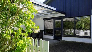 En bygning med en overdækket terrasse, et hvidt stakhegn og udsigt til grøn natur gennem vinduerne.
