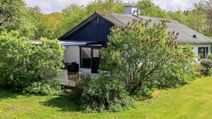 Et lille, etetages hus med et mørkeblåt tag og hvide vægge, omgivet af frodig vegetation og træer.
