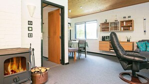 En hyggelig stue med en brændeovn, en behagelig læderstol, et spiseområde med bord og stole samt et køkken med hylder og en radio.