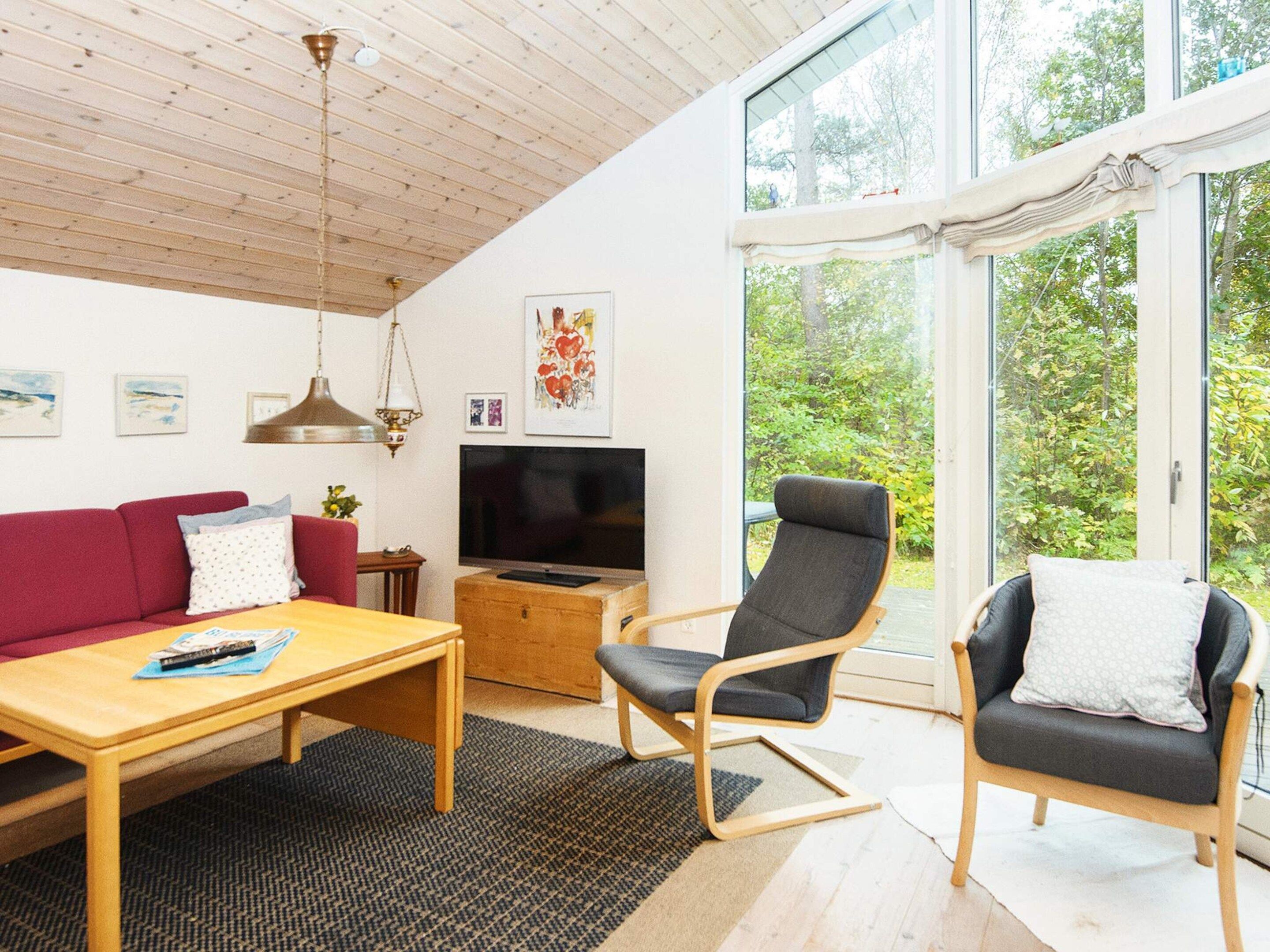 Ein modernes Wohnzimmer mit Holzbalkendecke, einem roten Sofa, einem Fernseher und einem großen Fenster mit Blick auf Bäume.