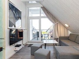 A modern loft bedroom with a large window, a flat-screen TV, and a cozy seating area with ottomans.