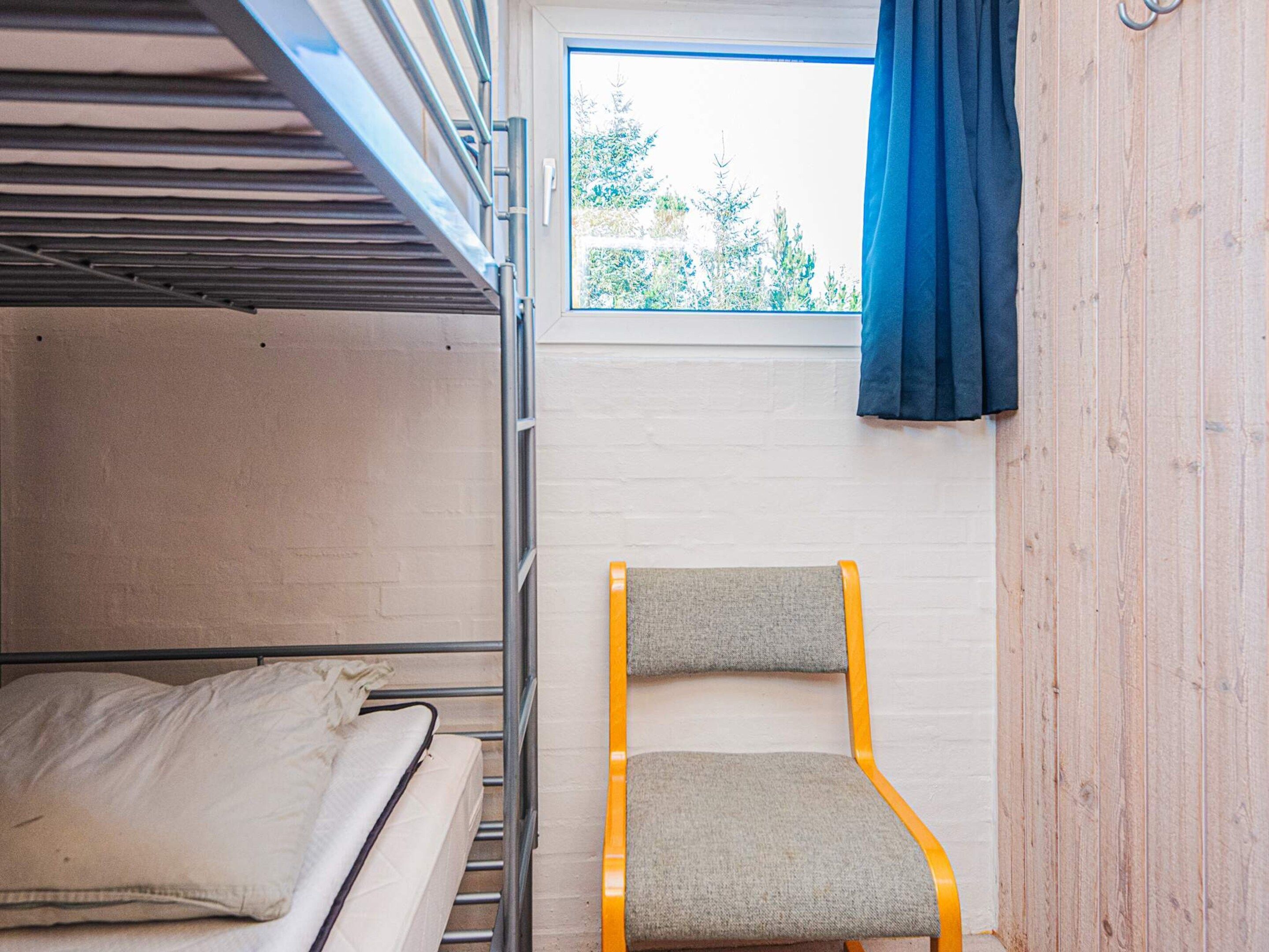 A bunk bed with a mattress and pillows, a chair with a cushion, and a window with a view of trees.