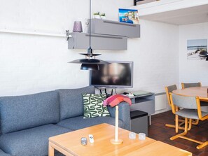 Uma sala de estar moderna com um sofá cinza, uma mesa de centro de madeira, uma TV e uma área de jantar com uma mesa redonda e cadeiras.