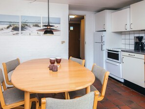 Uma sala de jantar com mesa de madeira e seis cadeiras, uma cozinha com armários brancos e uma obra de arte com tema praiano.