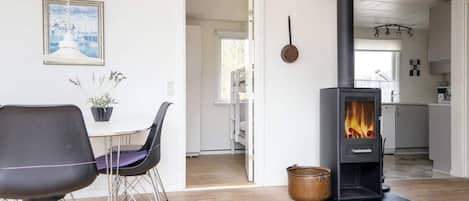 A modern living room with a black wood-burning stove, a dining table with chairs, and a view into the kitchen.