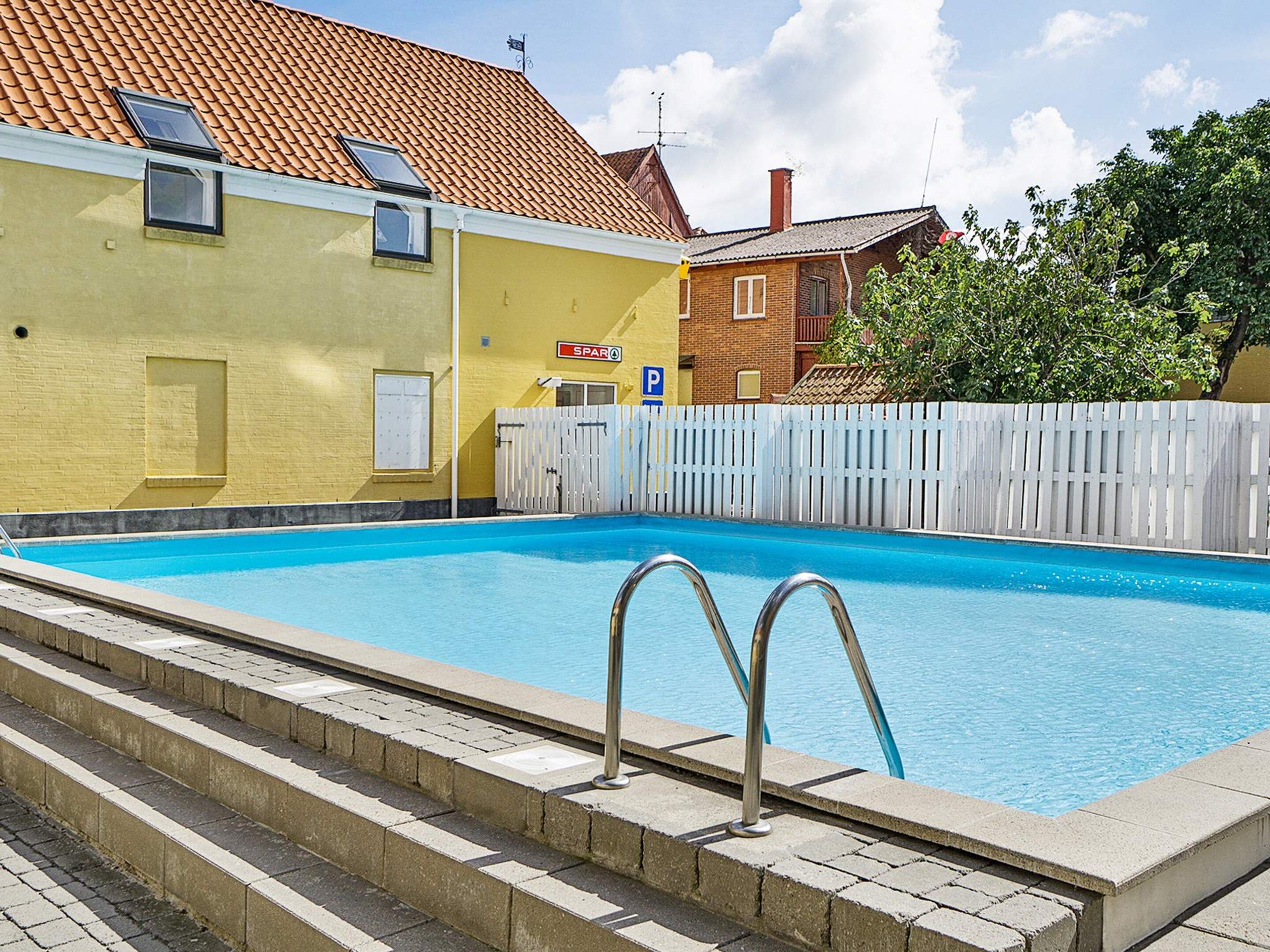 A swimming pool with steps leading into it, surrounded by a white fence, with a building in the background.