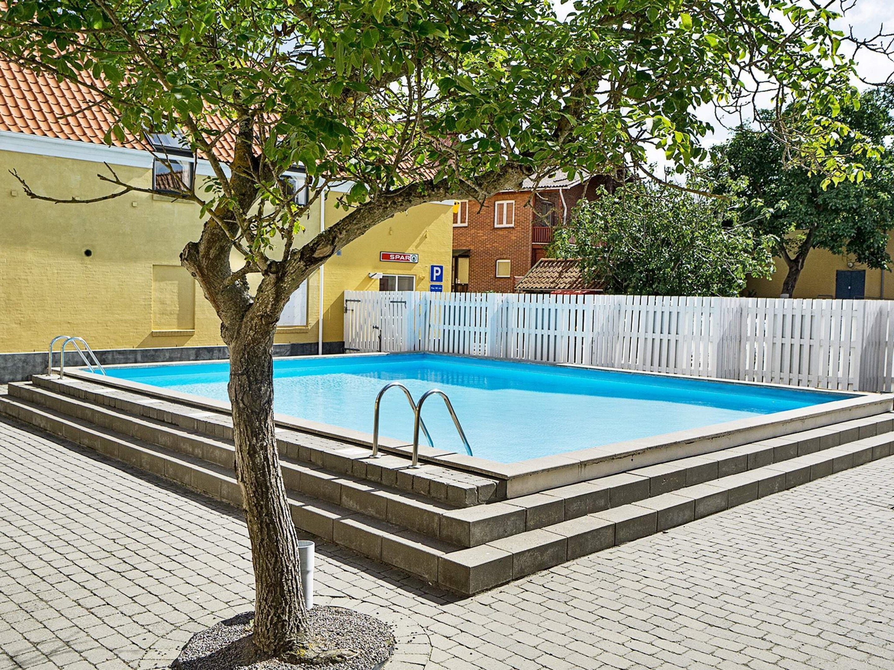A rectangular swimming pool with steps leading into it, surrounded by a white fence, and a tree in the foreground.