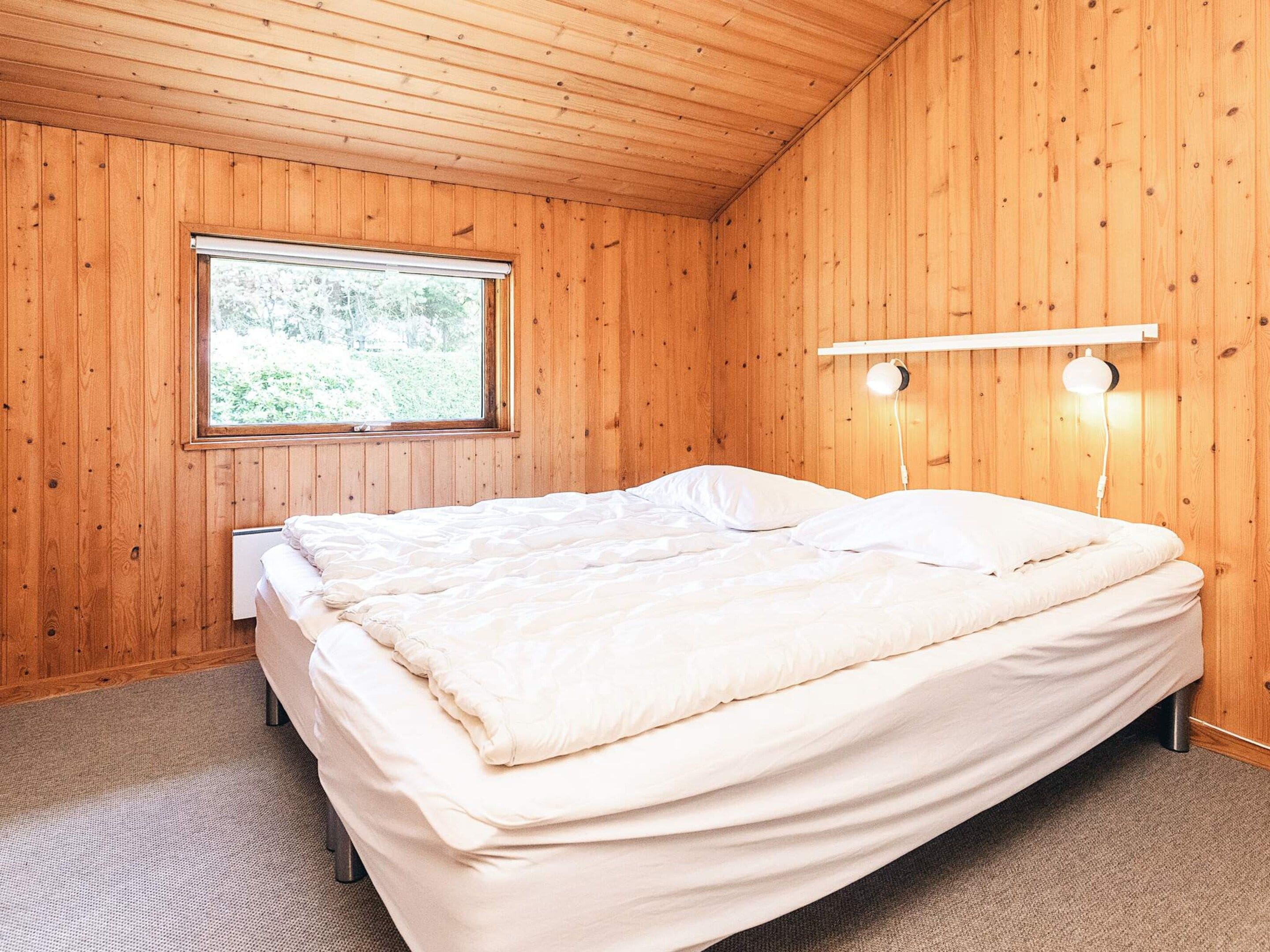 A room with wooden walls, a bed with white bedding, a window with a view of greenery, and two wall-mounted lights.