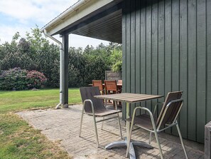 Un patio exterior con mesa y sillas, un área cubierta y un jardín con arbustos y árboles.