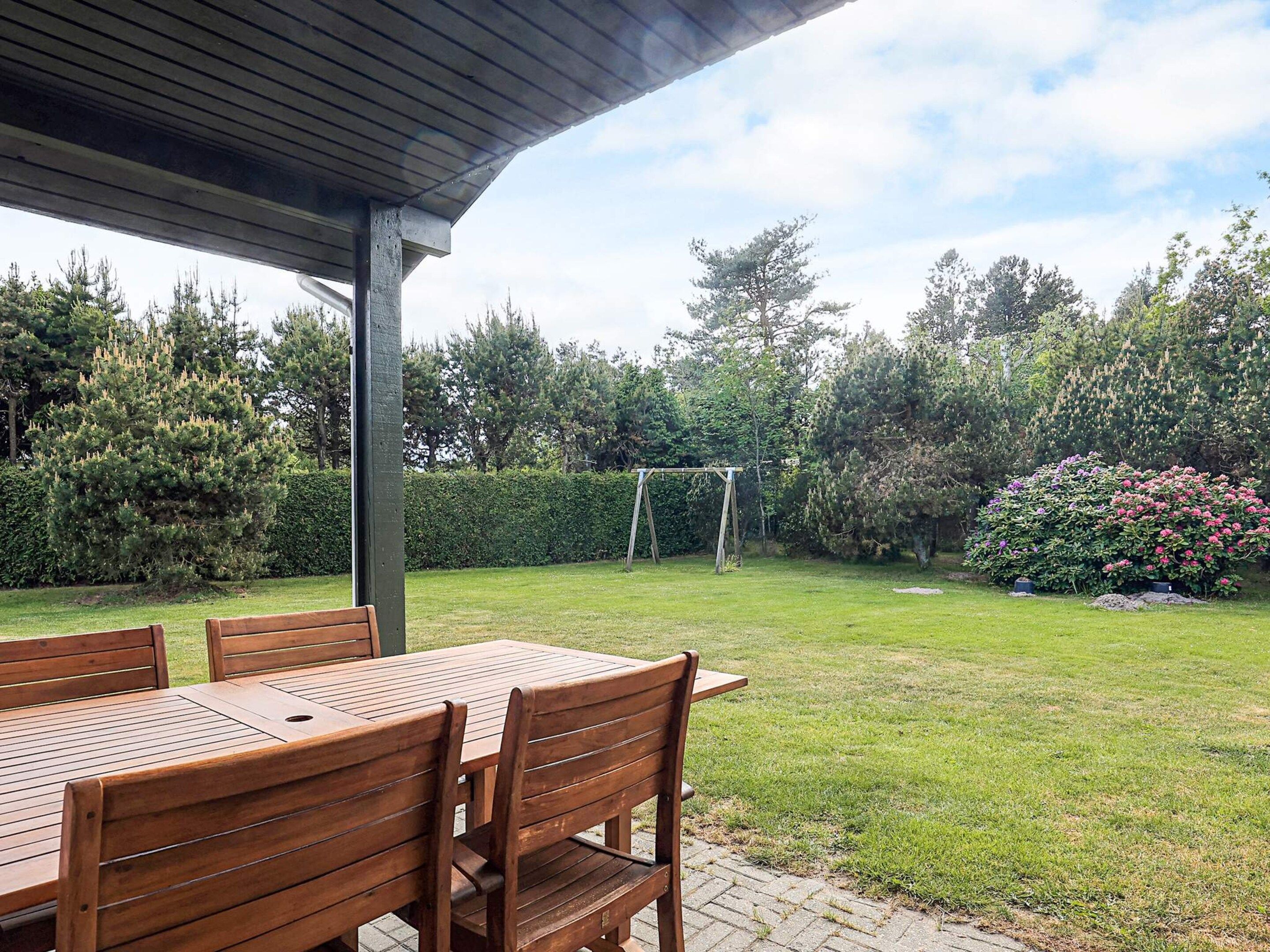 Une terrasse couverte avec des tables et des chaises en bois, une pelouse verdoyante, une balançoire en bois et une variété d’arbres et d’arbustes.