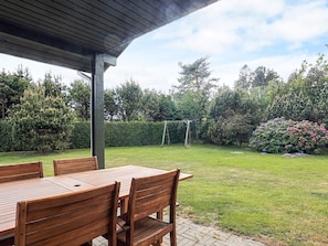 A covered patio with wooden tables and chairs, a grassy lawn, a wooden swing set, and a variety of trees and shrubs. - 4 Star Holiday Home in Vaeggerlose (Vaeggerlose)