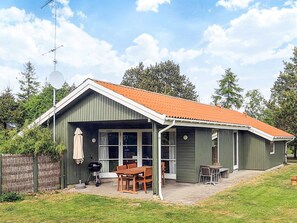 Ein eingeschossiges Haus mit überdachter Terrasse, Grill und Esstisch mit Stühlen.