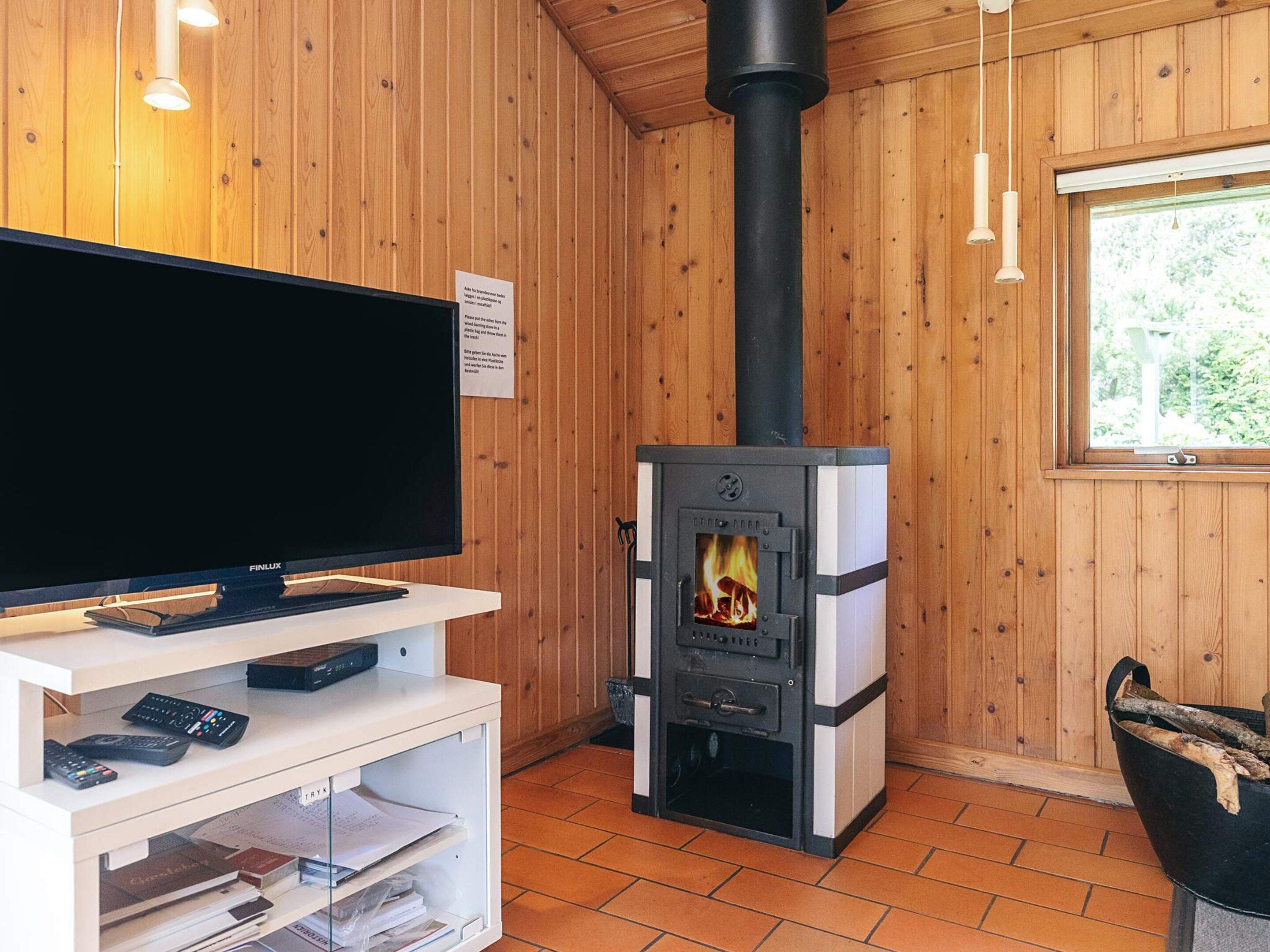 Une pièce chaleureuse avec un téléviseur posé sur un meuble blanc, une cheminée avec des bûches, des murs en bois et une fenêtre.