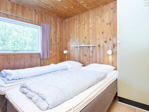 A bedroom with a wooden paneled wall, a window with a curtain, a bedside table with a lamp, and a bed with pillows and a duvet.