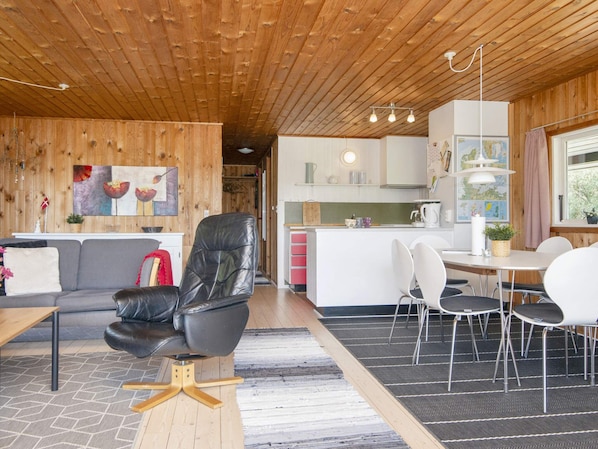 A modern interior with a wooden ceiling, a dining area with white chairs, a black leather sofa, and a kitchen area with white cabinets.