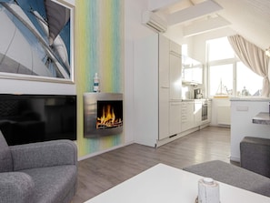 A modern living room with a fireplace, a kitchen in the background, and a large window with curtains. - Holiday Home in Wendtorf (Wendtorf)