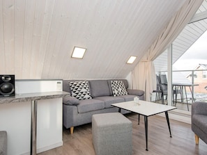 A modern loft-style room with a grey sofa, a small white table, and a kitchen area with a microwave and speaker.