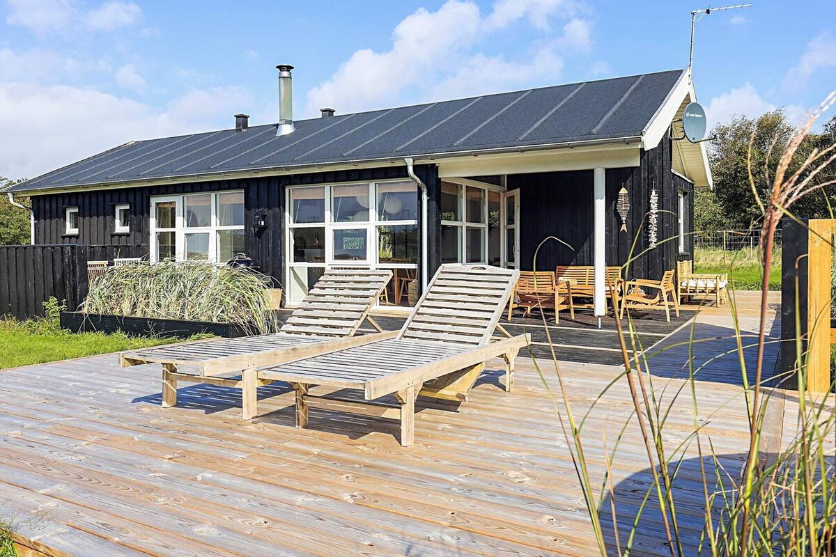 A wooden deck with lounge chairs, a wooden table, and a black building with large windows and a chimney.
